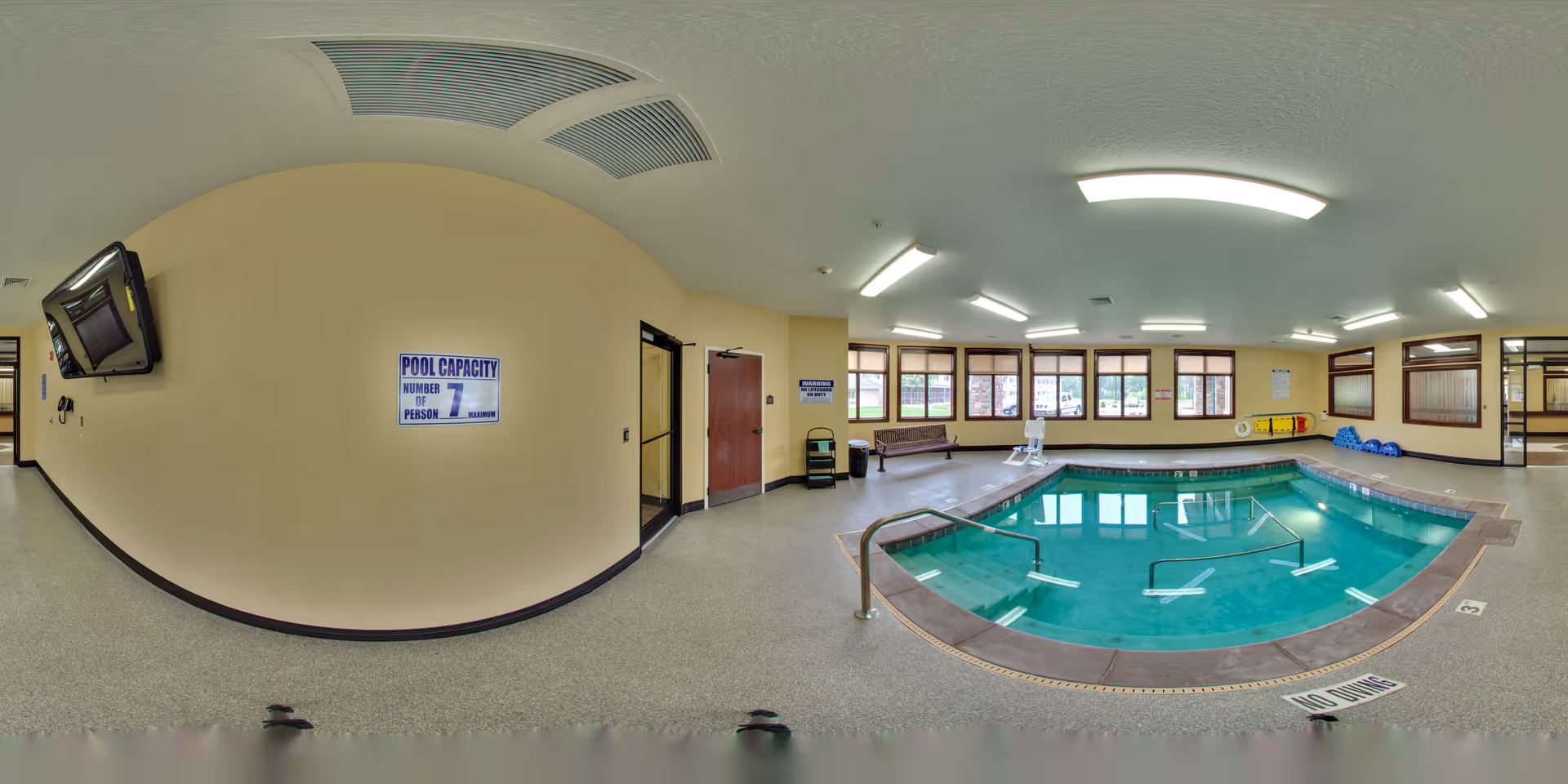 Indoor pool area with a small rectangular pool surrounded by a tiled edge and handrails. The room has beige walls, multiple windows letting in natural light, and ceiling lights. There are benches, pool safety equipment, and signs on the walls including one indicating a pool capacity of 7 people. The floor is covered with a speckled non-slip surface.