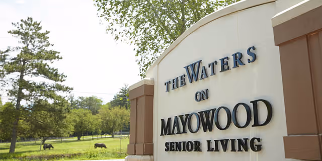 Outdoor view of a large sign for The Waters on Mayowood Senior Living facility, with trees and grassy area in the background under a clear sky.