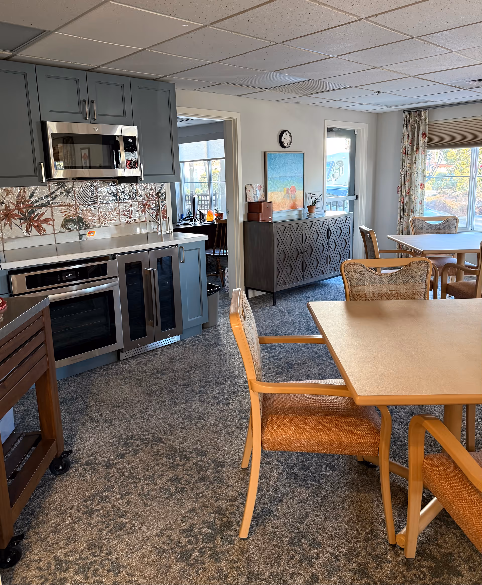 A senior living facility dining area with wooden tables and chairs, adjacent to a kitchen area featuring blue cabinets, a microwave, an oven, and a wine cooler. The room has carpeted flooring, a sideboard with decorative items, a clock on the wall, and large windows with floral curtains letting in natural light.