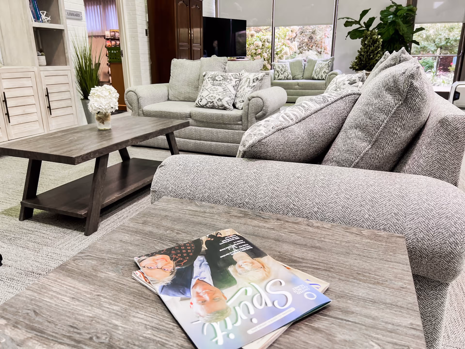 A cozy living room area with gray upholstered sofas and armchairs, a wooden coffee table with a vase of white flowers, and a magazine on a side table. There is a large window with a view of greenery outside, and a TV and bookshelf are visible in the background.