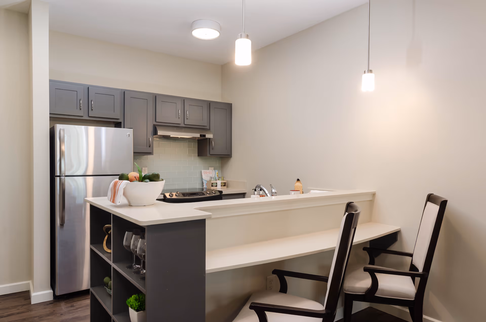 Modern kitchen area with gray cabinets, a stainless steel refrigerator, a stove, and a white countertop with a built-in sink. Two cushioned chairs are placed at the counter, and pendant lights hang from the ceiling above the counter. A bowl with fruit and a towel are on the counter.