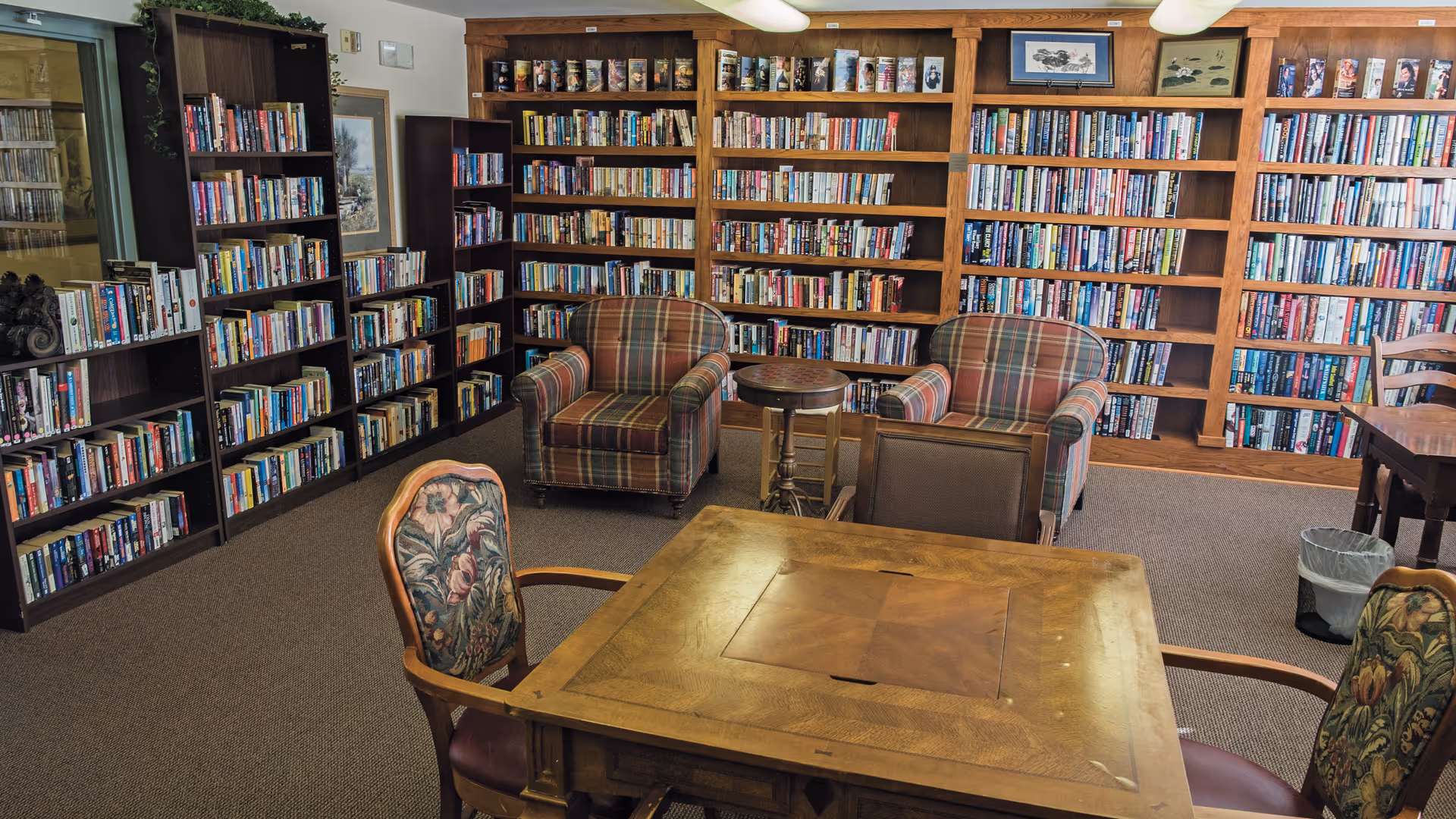 Library-style common room with floor-to-ceiling bookshelves, plaid armchairs, and a wooden table with chairs.