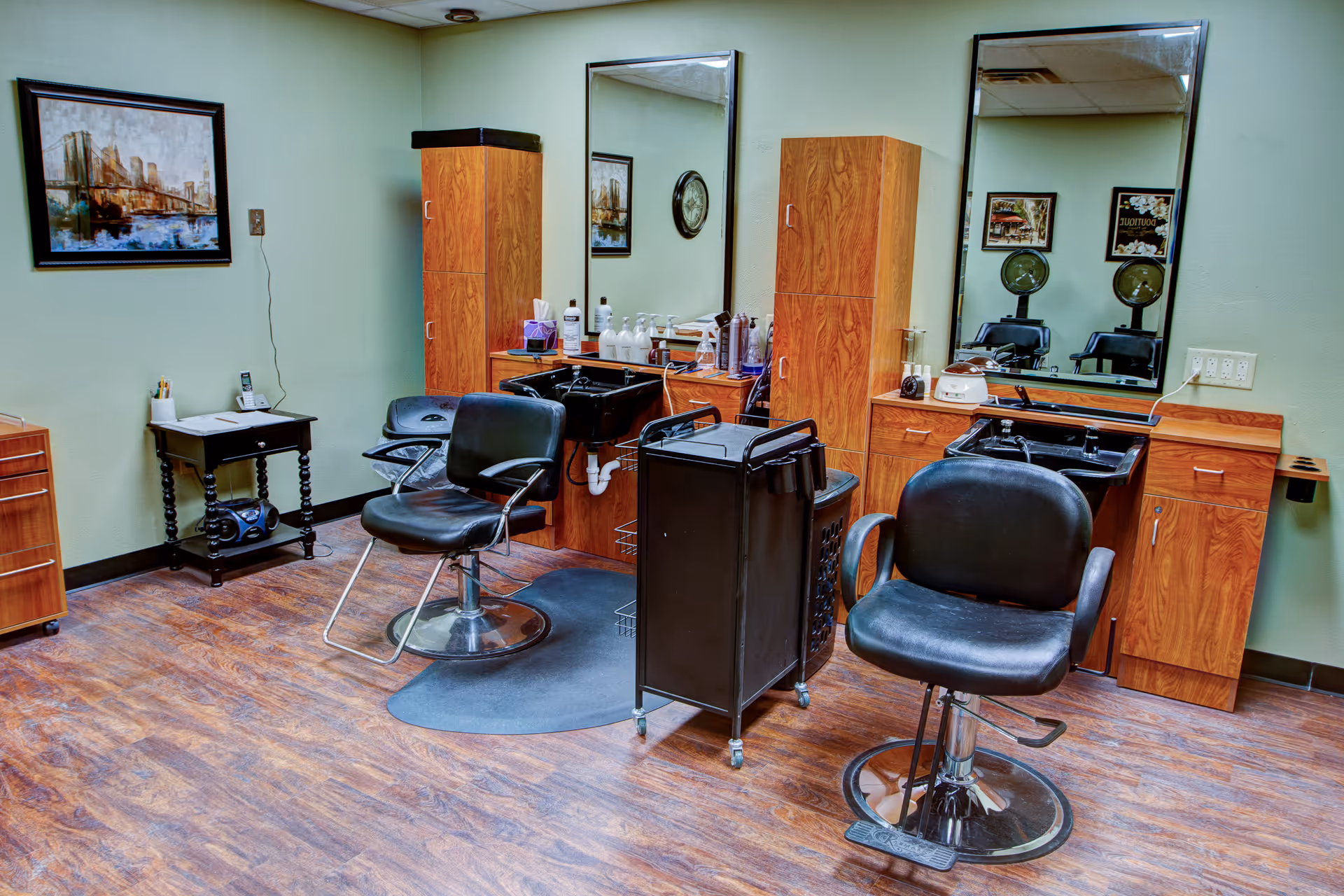 Interior view of a salon area in a senior living facility with two black salon chairs in front of two large mirrors mounted on a light green wall. Below the mirrors are wooden cabinets and black sinks for hair washing. Various hair care products are placed on the counter. The floor has a wood-like finish, and there is a small black table with a phone and a radio on it. The room is well-lit and neatly organized.