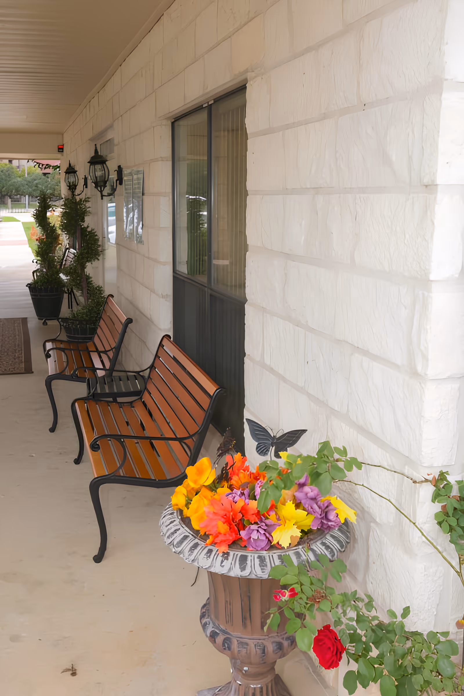 Covered outdoor walkway with wooden benches along a stone wall, decorative wall-mounted lanterns, potted plants, and a large planter filled with colorful artificial flowers and a butterfly decoration.