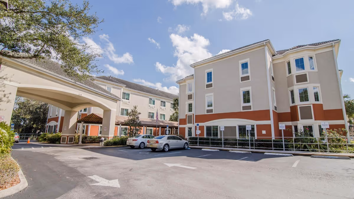 Exterior view of a multi-story senior living facility building with beige and brown walls, several windows, and a covered entrance. There are a few parked cars in the parking lot in front of the building, and trees and shrubs around the area under a partly cloudy blue sky.