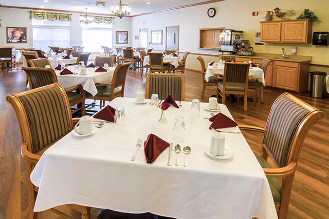 A bright dining room in a senior living facility with multiple tables covered in white tablecloths, each set with cups, glasses, silverware, and folded burgundy napkins. The room features wooden chairs with striped cushions, large windows with valances, and a beverage station with cabinets and a coffee machine in the background.