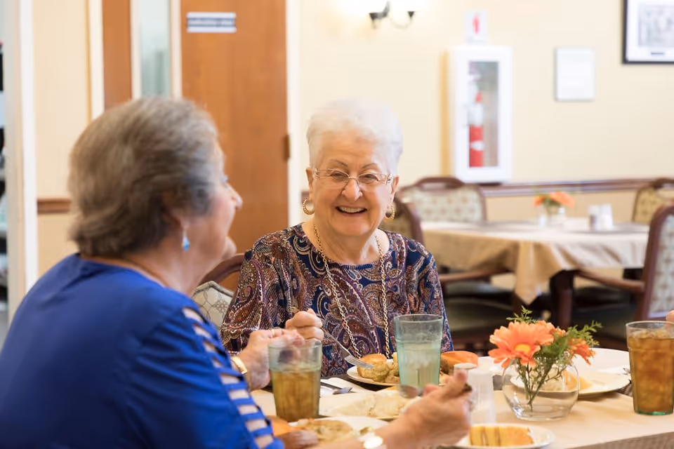 Two elderly women sitting at a dining table in a senior living facility, enjoying a meal together and smiling. The table is set with plates of food, glasses of iced tea, and a small vase with orange flowers. The background shows other tables with beige tablecloths and chairs in a well-lit dining room.