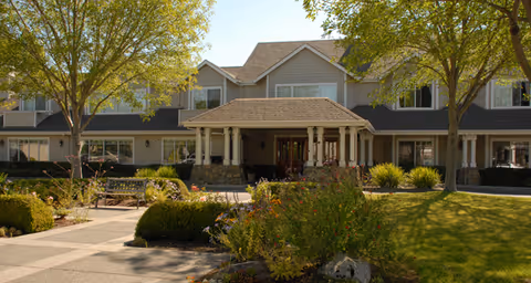 Front exterior of a two-story senior living building with a covered entry, landscaped courtyard, and trees.