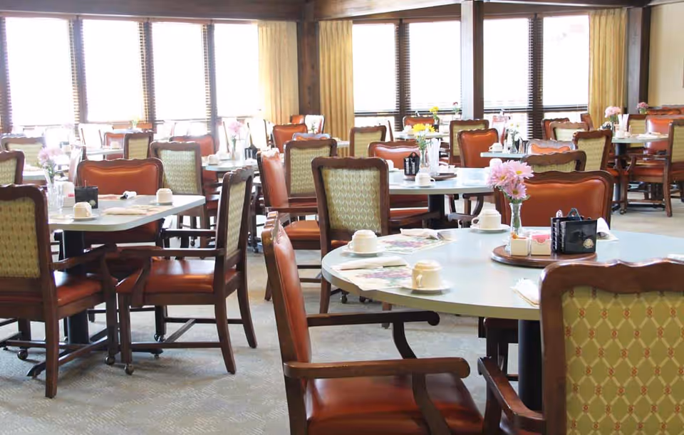 A bright dining room with multiple tables and chairs arranged neatly. Each table has cups, napkins, and small flower vases with pink and yellow flowers. Large windows with blinds and curtains allow natural light to fill the room.