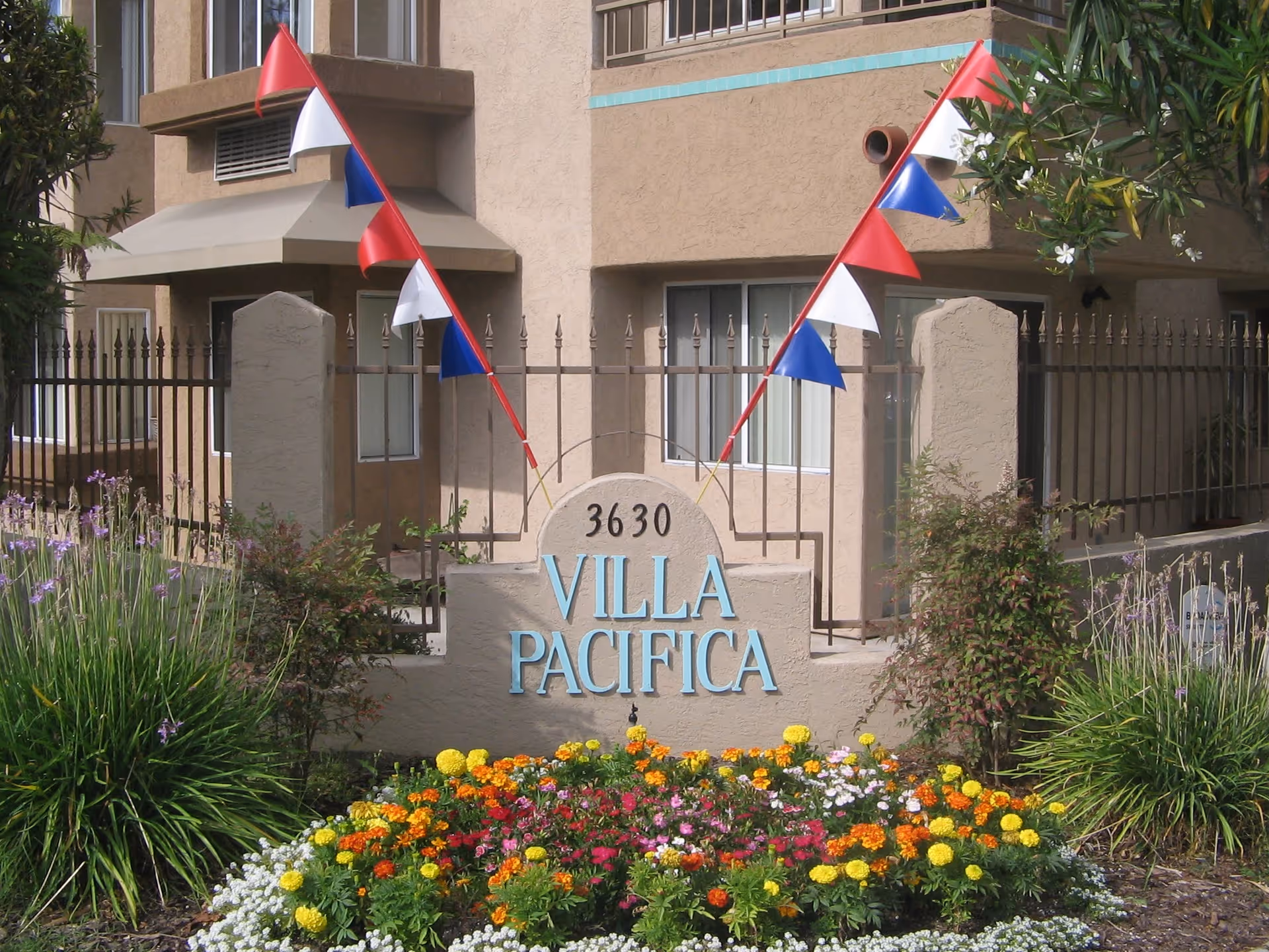Entrance sign reading "Villa Pacifica" with the address 3630 in front of a stucco building, pennant flags, and a colorful flower bed.