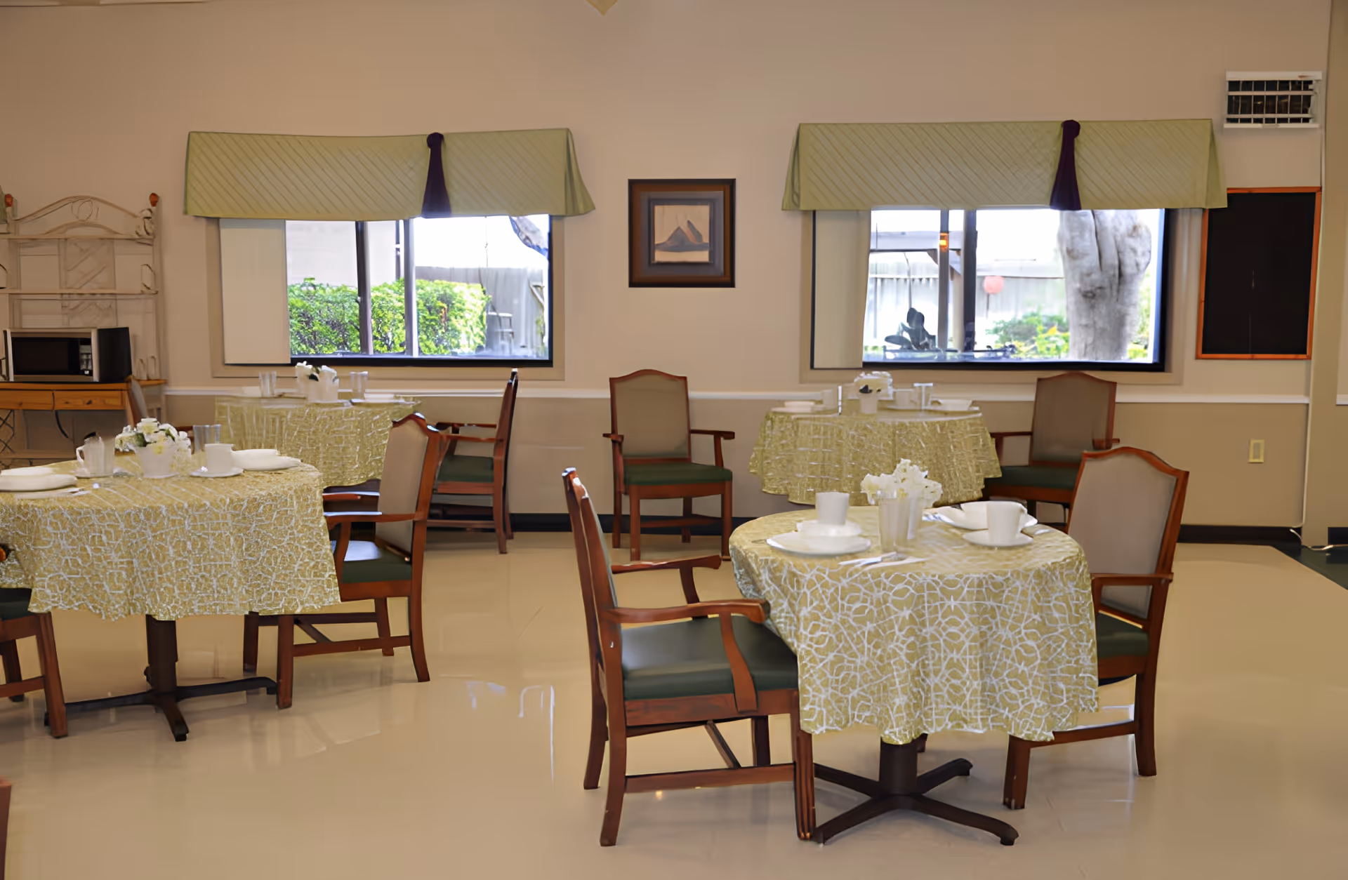 A dining room with several round tables covered with yellow patterned tablecloths. Each table is set with white plates, cups, and utensils, and surrounded by wooden chairs with green cushions. Two large windows with green valances let in natural light, and a framed picture hangs on the beige wall between the windows. A microwave is visible on a wooden shelf in the corner.