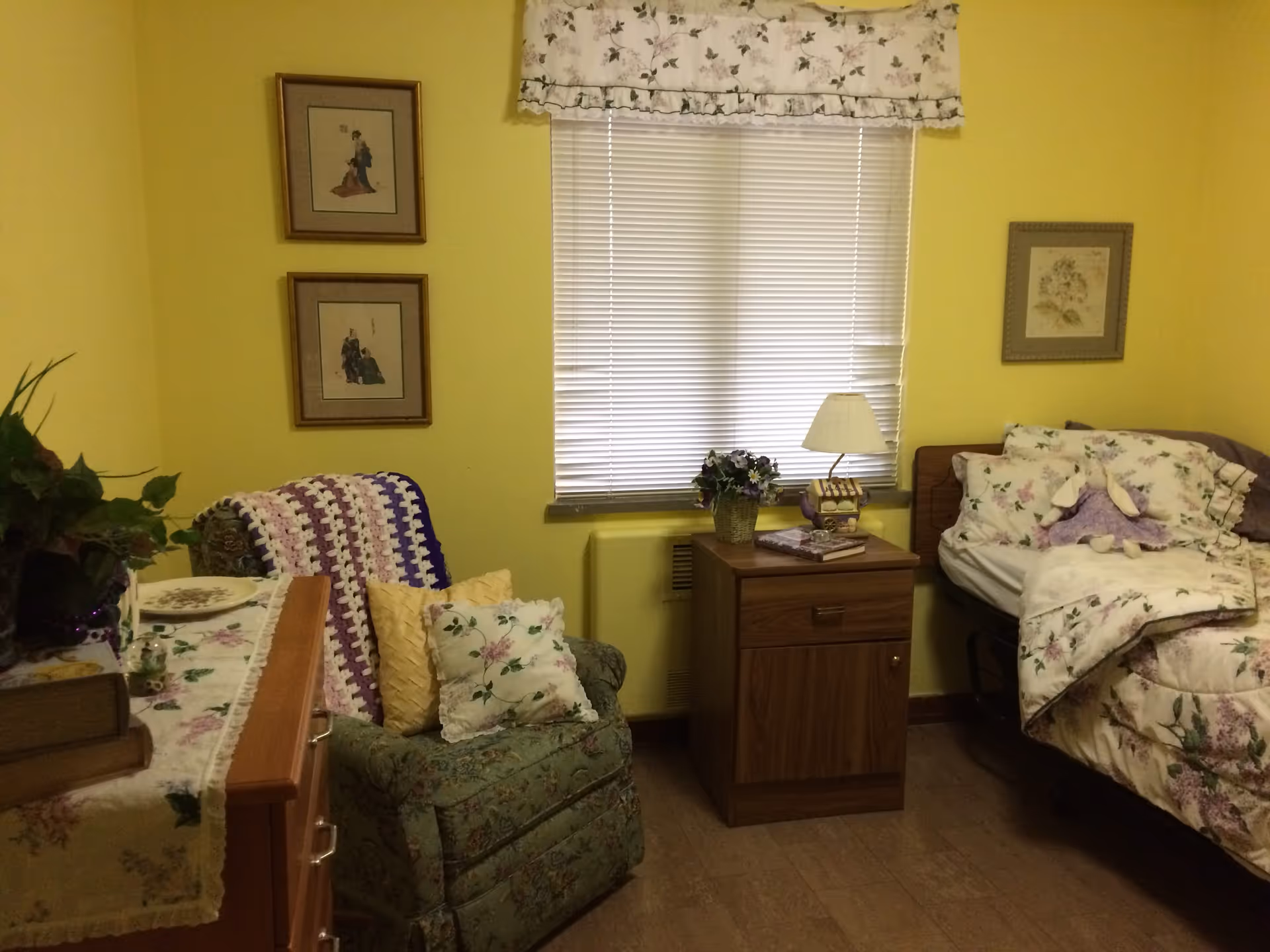 A cozy bedroom with yellow walls featuring a bed with floral bedding and a stuffed bunny, a wooden nightstand with a lamp and flowers, a floral armchair with pillows and a crocheted blanket, a dresser with decorative items, and framed artwork on the walls.