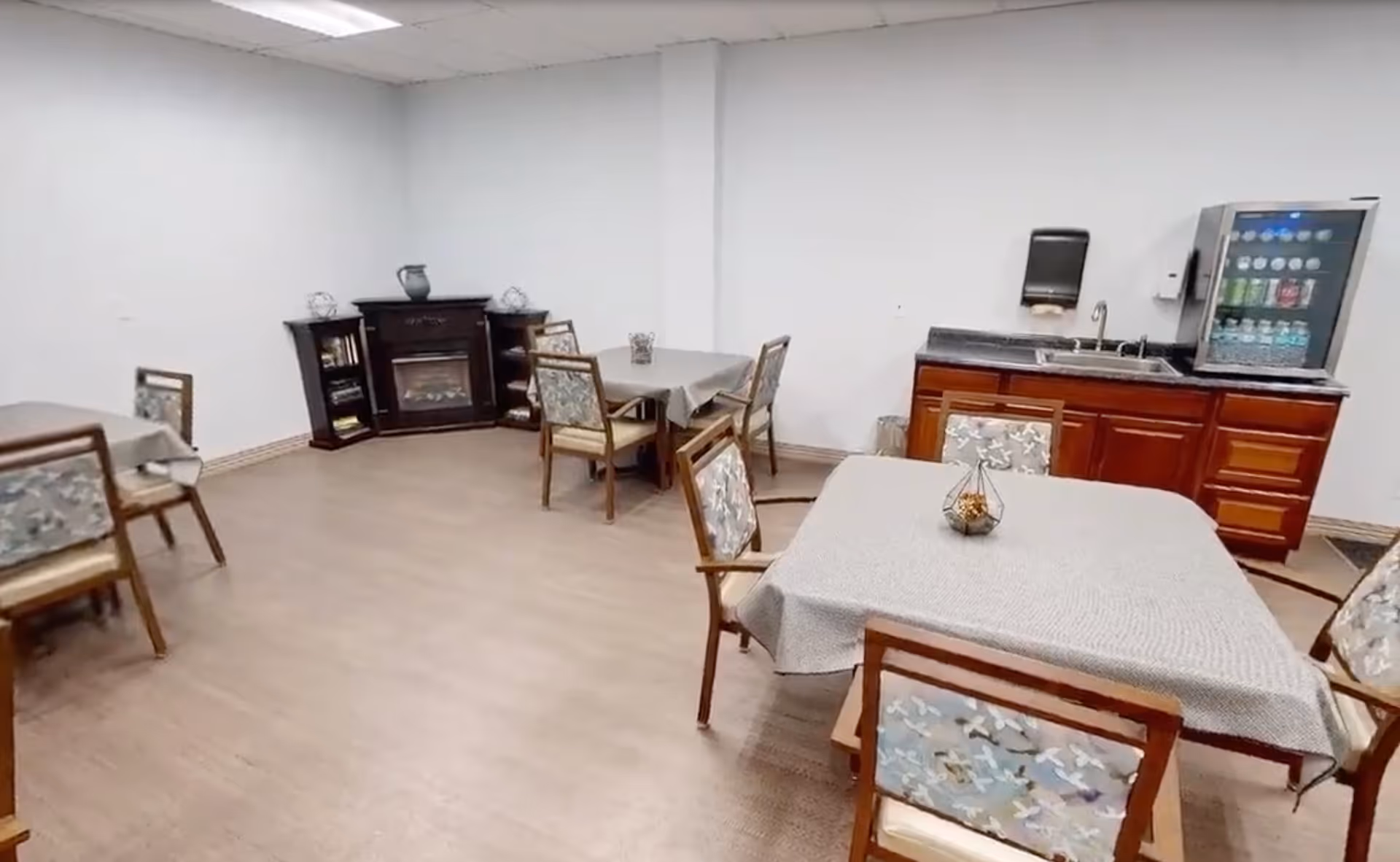 A dining area with several tables covered with light gray tablecloths and wooden chairs with patterned cushions. In the background, there is a dark wooden electric fireplace with shelves on either side holding decorative items. To the right, there is a countertop with a sink, a paper towel dispenser, and a beverage vending machine. The room has light-colored walls and a wood-look floor.