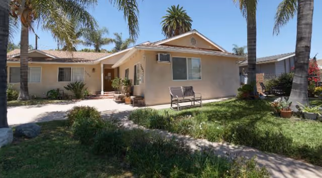 Single-story beige senior care building with front lawn, palm trees, and a bench.