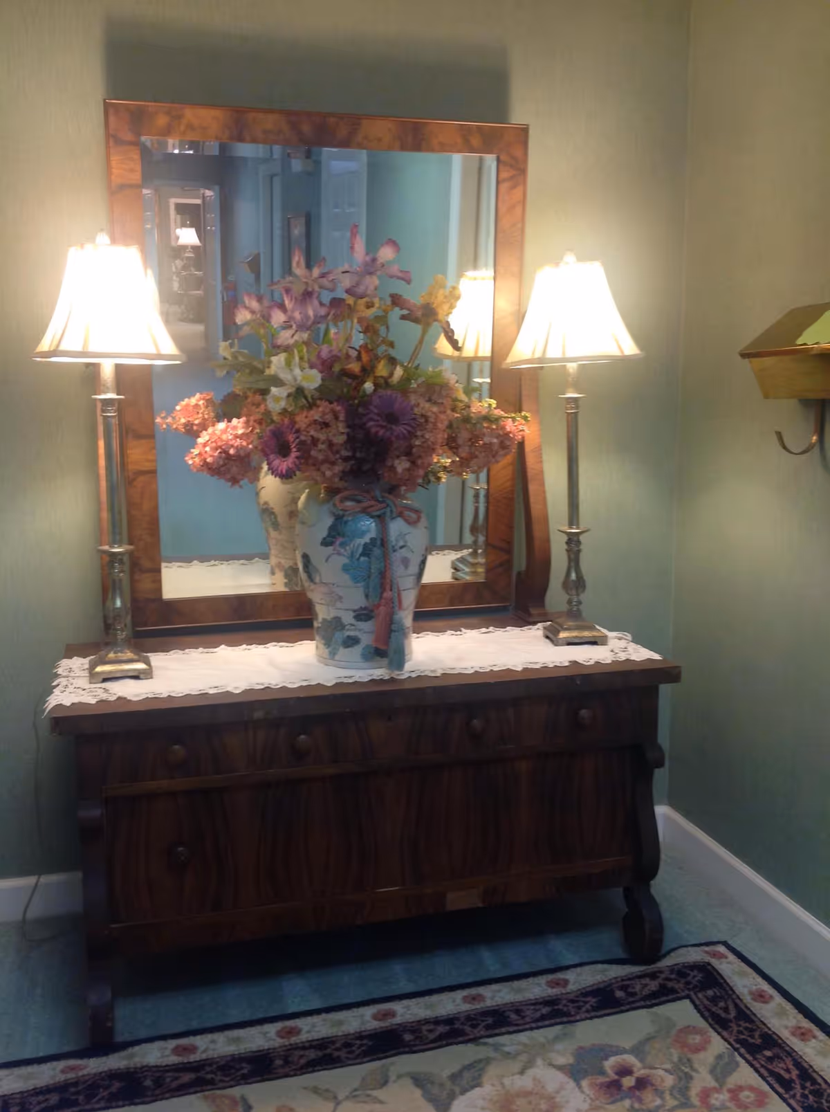 A wooden dresser with a large rectangular mirror above it, flanked by two lit table lamps with white shades. On the dresser is a decorative vase filled with a colorful arrangement of flowers. The dresser is placed against a green wall, and a floral-patterned rug is partially visible on the floor.