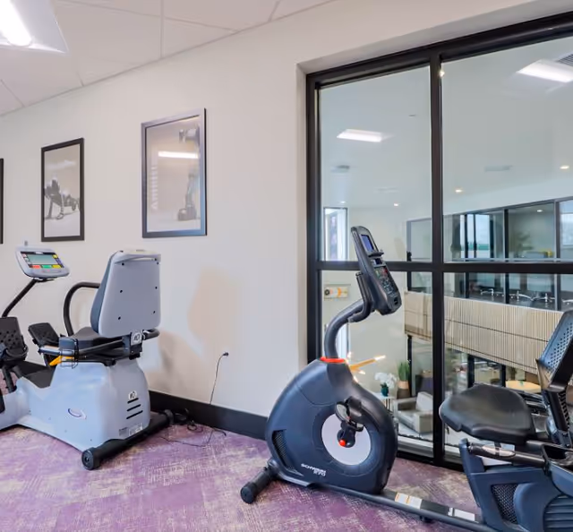 Indoor exercise room with stationary bikes placed on a purple carpeted floor. The room has white walls with framed motivational posters and a large window looking into another interior space with seating and plants.