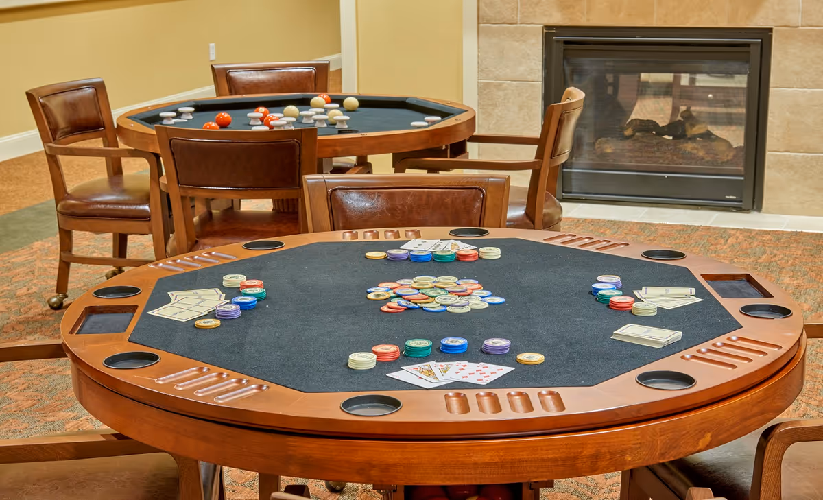 Two octagonal wooden game tables with black felt tops in a room. One table is set up for poker with playing cards and poker chips, and the other table has billiard balls arranged on it. Brown leather chairs surround the tables, and there is a fireplace with a glass front in the background.