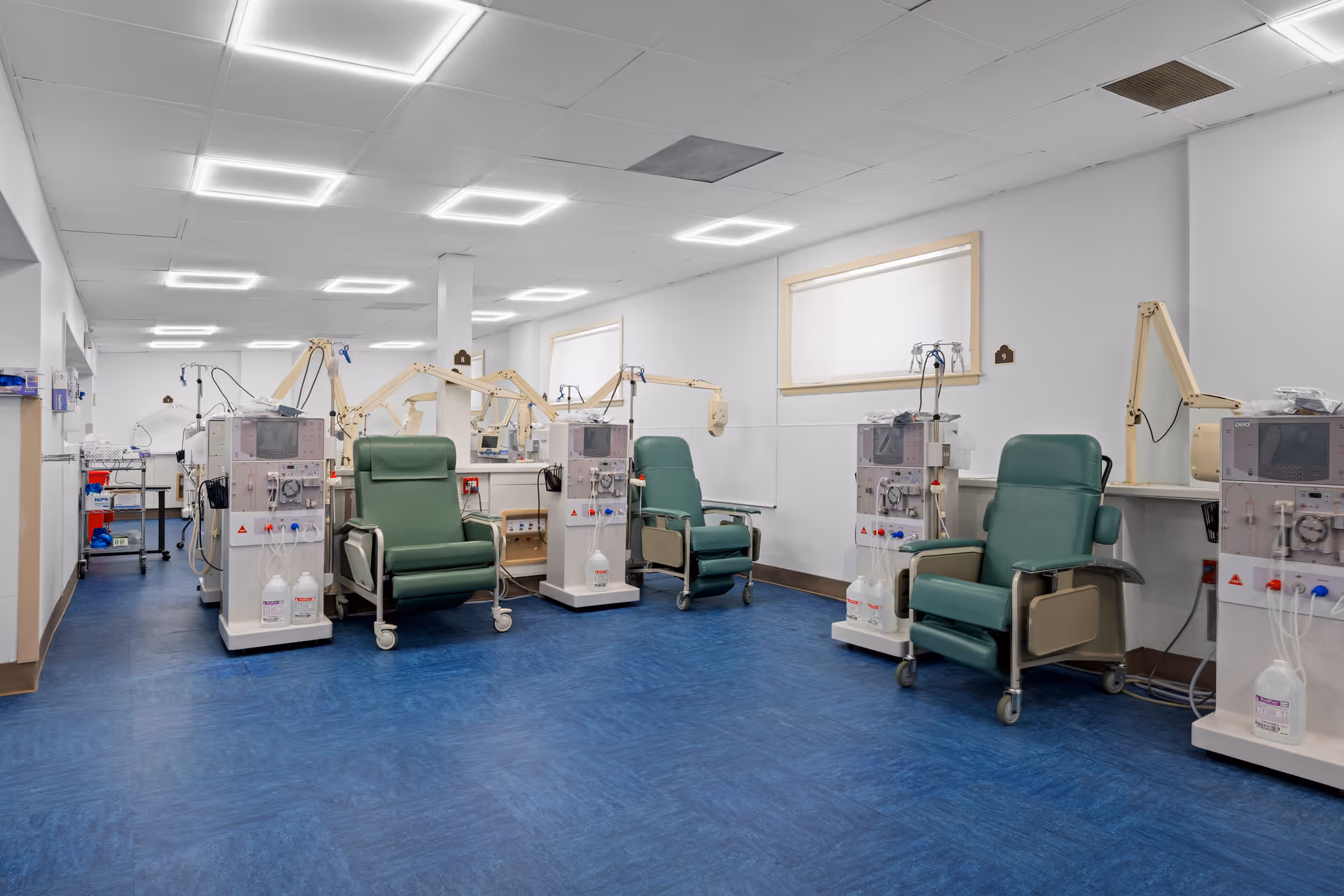 A spacious clinical treatment room with multiple green reclining dialysis chairs and dialysis machines arranged along the walls.