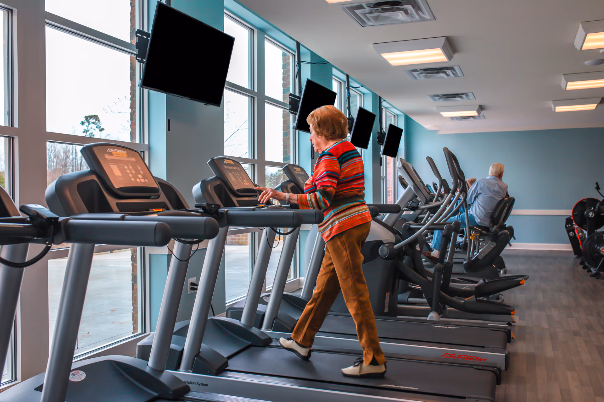 An elderly woman in a colorful striped sweater and brown pants walking on a treadmill in a fitness room with large windows and multiple treadmills and exercise bikes. A man is seated on one of the exercise bikes in the background. Several wall-mounted televisions are visible above the equipment.