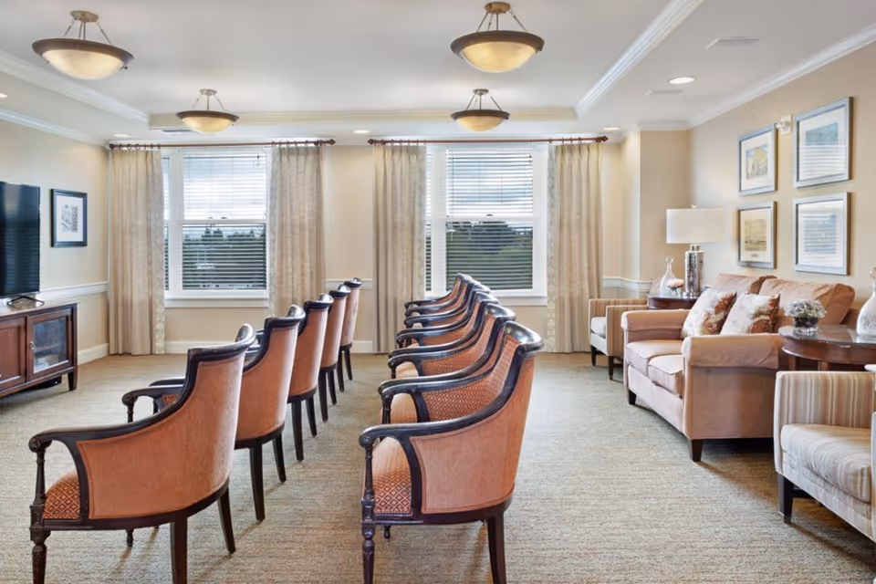 Well-lit communal sitting room with two rows of upholstered armchairs facing forward and a sofa seating area by windows.