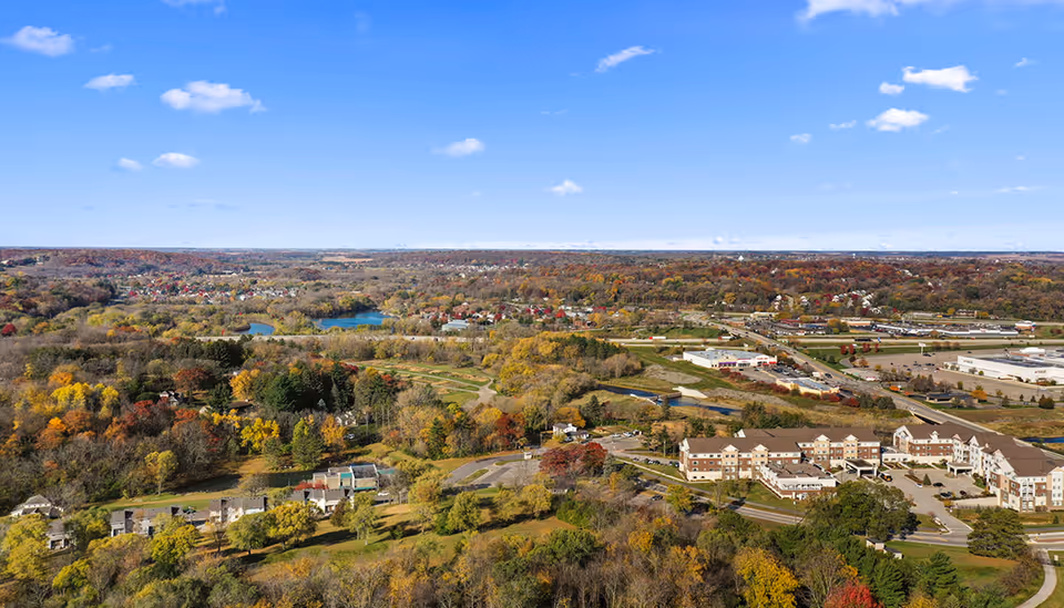 Aerial view of The Waters on Mayowood campus amid fall-colored trees, nearby lakes, roads and buildings under a blue sky.