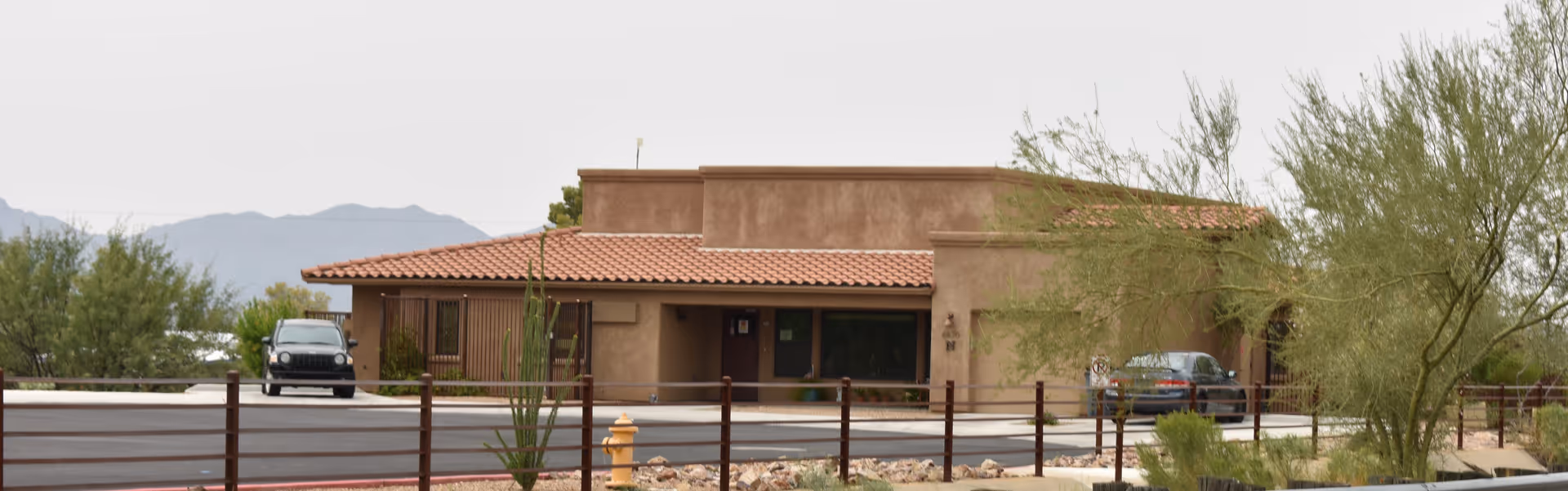 Exterior view of a single-story building with a flat roof and terracotta tiles, surrounded by desert landscaping including small trees and shrubs. Two cars are parked near the building, and mountains are visible in the background under a cloudy sky.