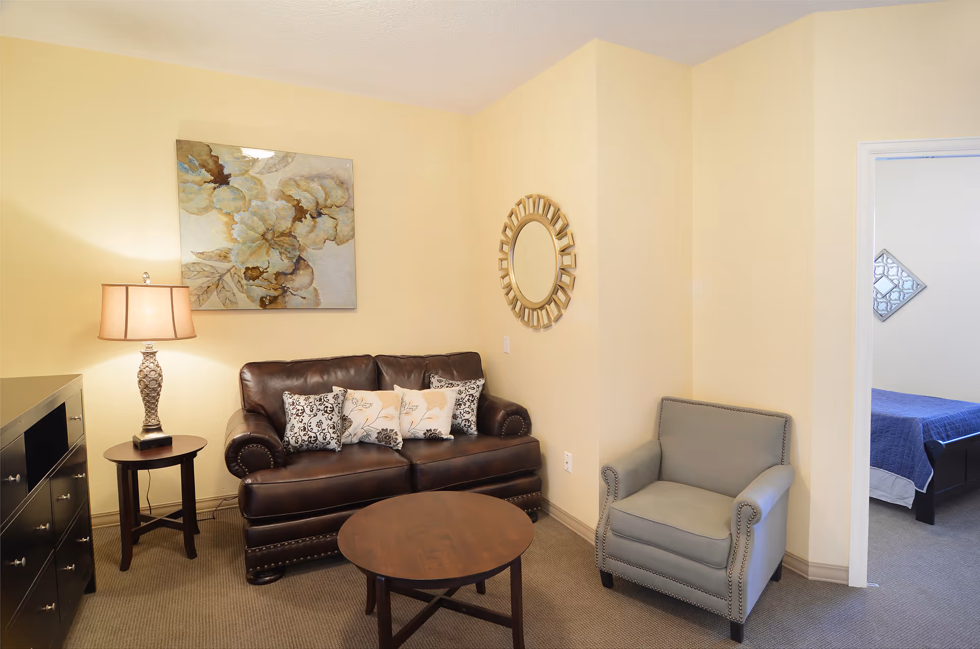 A cozy living room area with a dark brown leather loveseat adorned with decorative pillows, a round wooden coffee table, a light gray armchair, a side table with a lamp, and a floral painting on the wall. A round mirror hangs on the adjacent wall, and an open doorway reveals a bedroom with a blue bedspread.