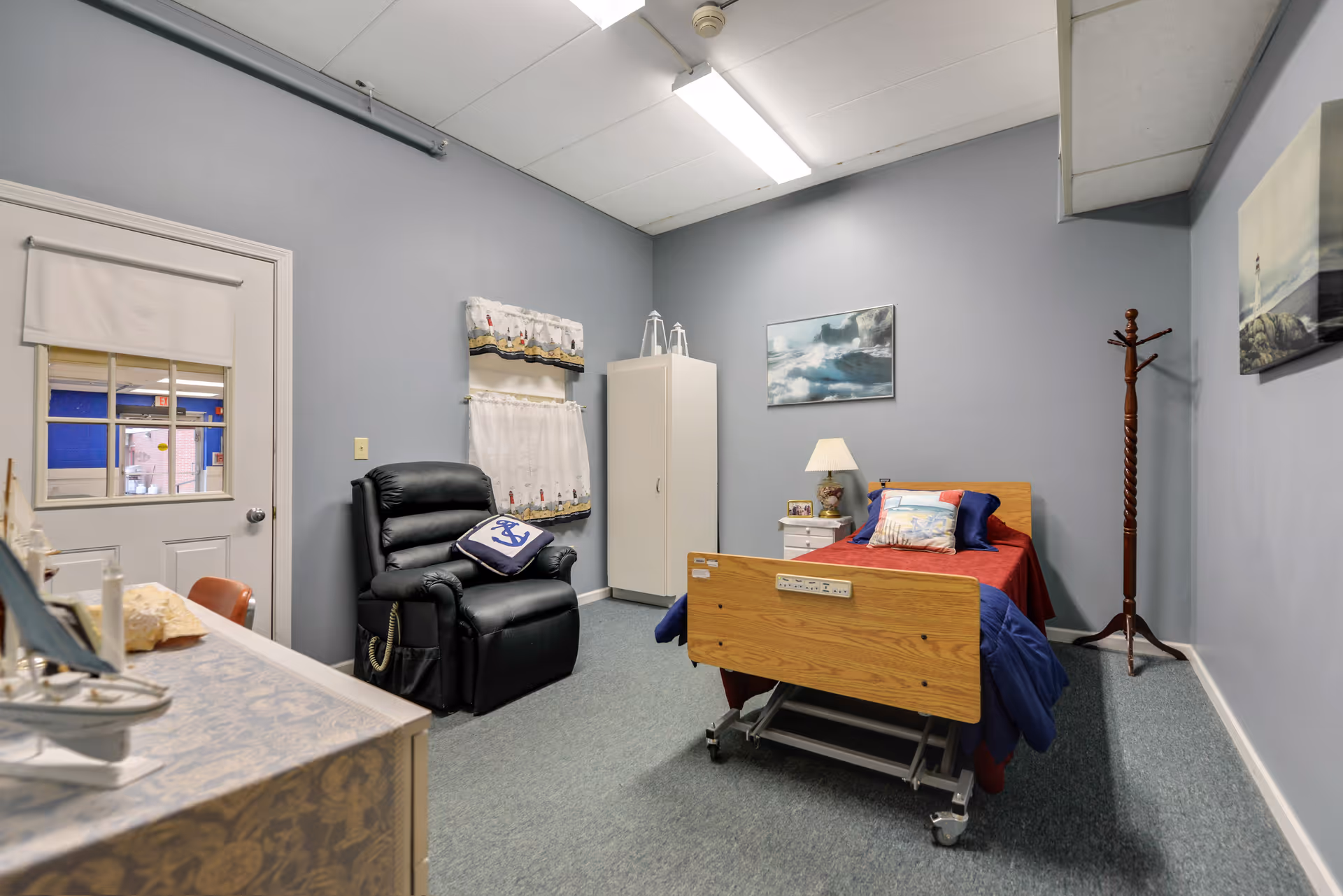 A small, cozy room with a single hospital-style bed covered with a red and blue blanket. There is a black recliner chair with a nautical-themed pillow, a white cabinet, a wooden coat rack, and a nightstand with a lamp and small decorative items. The walls are painted light blue and decorated with ocean-themed paintings and curtains with a lighthouse pattern. A door with a window is visible on the left side.