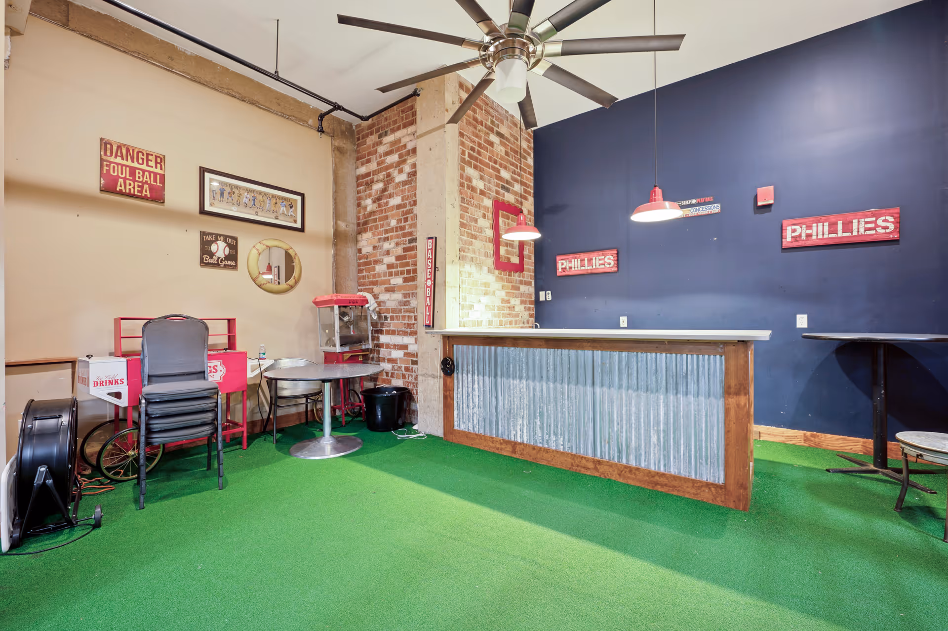 An indoor recreational area with a green carpet floor resembling artificial turf, a ceiling fan, and a bar counter with corrugated metal front. The walls are decorated with baseball-themed signs including 'PHILLIES' and 'DANGER FOUL BALL AREA'. There is a popcorn machine, stacked chairs, and small round tables in the room.