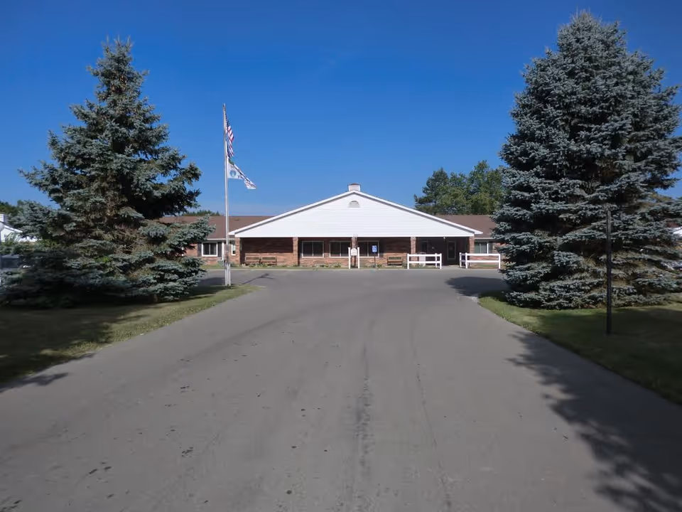 Front exterior view of Medilodge of Sterling building with a paved driveway leading up to the entrance, flanked by two large evergreen trees and flagpoles displaying the American flag and other flags under a clear blue sky.