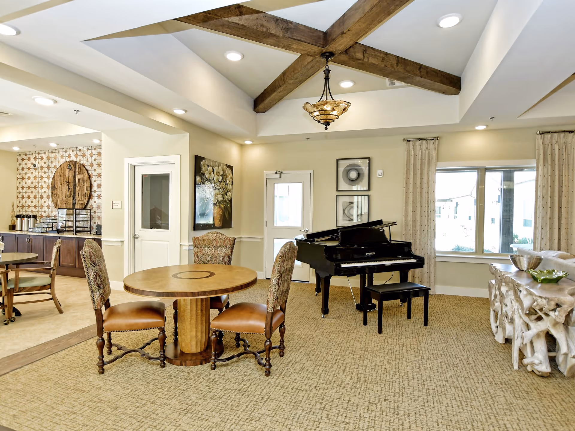 Bright senior living common room with a round table and chairs, a grand piano, and exposed ceiling beams.
