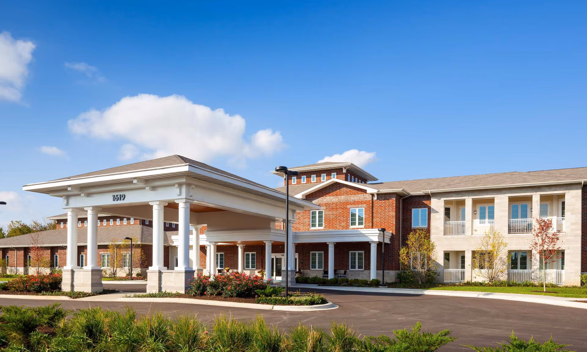 Exterior view of HarborChase of Naperville, a senior living facility with a large covered entrance supported by white columns, red brick and light stone facade, surrounded by landscaped greenery and a clear blue sky.