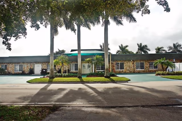 Front exterior view of Margate Assisted Living facility with a single-story stone building, green awning over the entrance, palm trees, and a paved driveway.