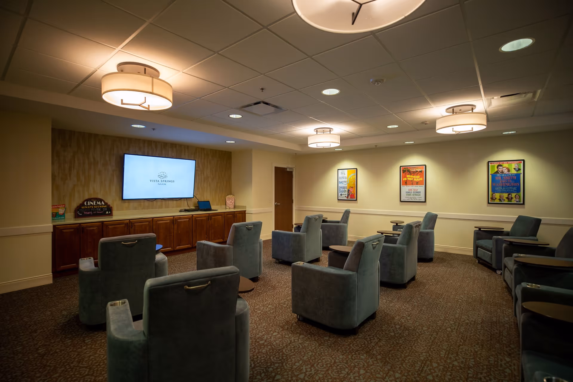 A senior living community media room with upholstered recliner chairs arranged facing a wall-mounted TV and framed posters on the walls.