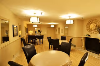 Interior view of a common area in a senior living facility with round tables and black chairs, a piano in the background, warm lighting from ceiling fixtures, a large decorative clock on the wall, and a fireplace.