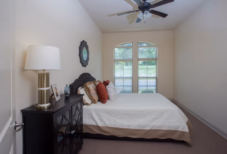 Bedroom with a bed dressed in a white quilt and pillows, a dark nightstand with a lamp, and a large window with blinds.