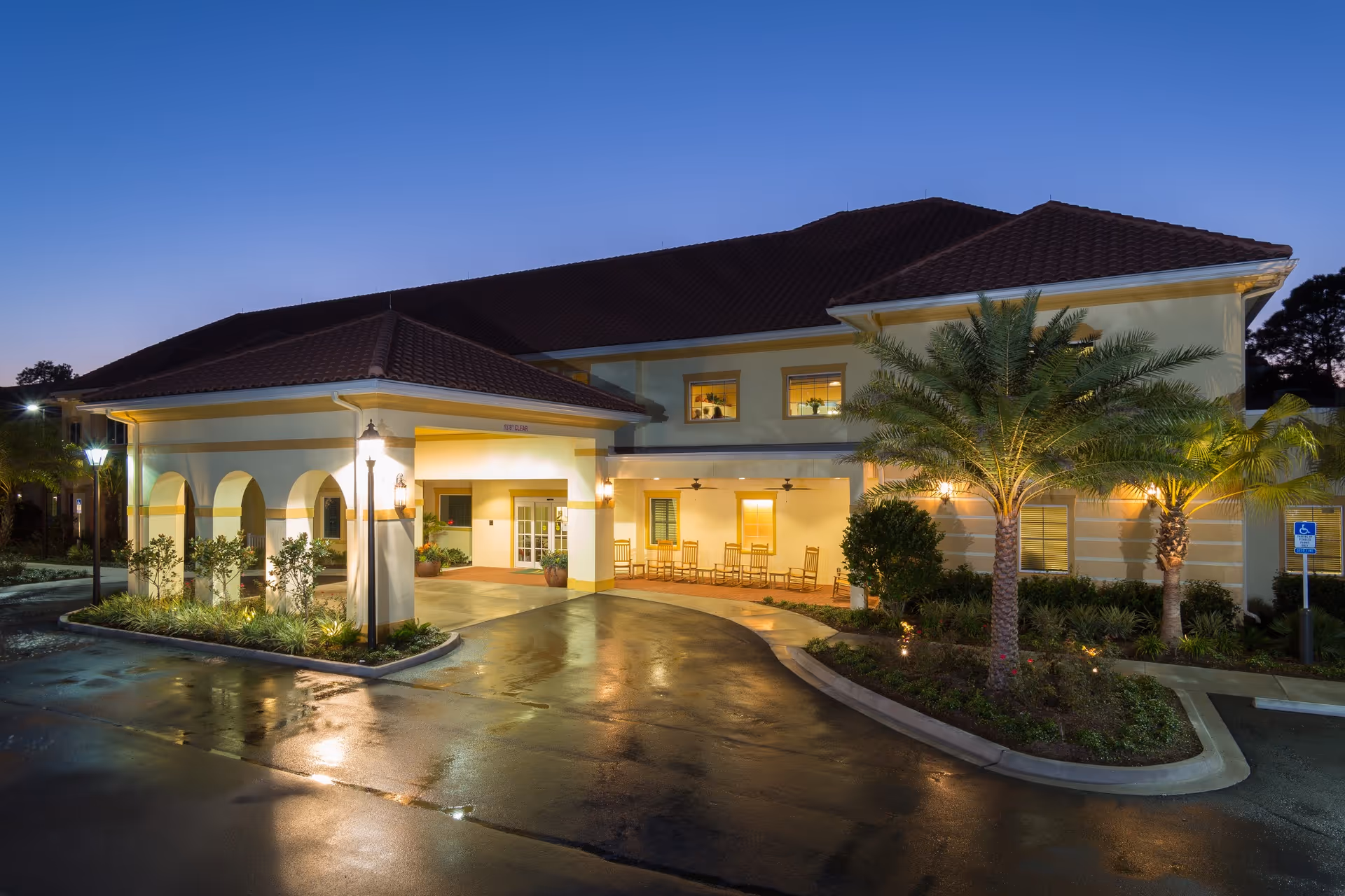 Front entrance of a two-story senior living building at dusk with a covered drop-off, rocking chairs, palm trees, and a wet driveway.