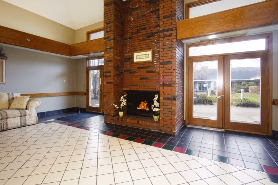 Bright lobby area with a central brick fireplace, tiled floor, a sofa to the left, and glass double doors leading outside.