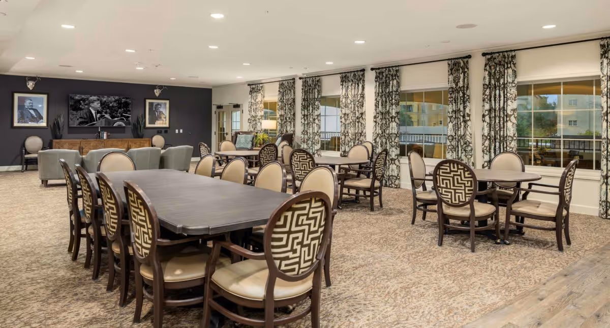 Spacious communal dining and activity room with multiple tables and patterned chairs, large windows with drapes, and a seating area with a TV.