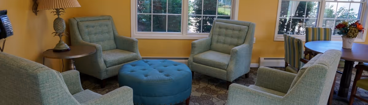 A cozy living room area with four light blue upholstered armchairs arranged around a round blue tufted ottoman. The room has yellow walls, large windows letting in natural light, a small round wooden side table with a pineapple-shaped lamp, and a round dining table with striped chairs and a floral centerpiece in the background.