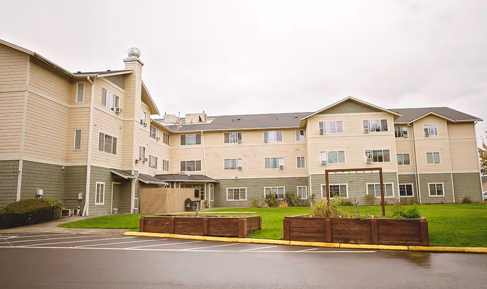 Exterior view of a three-story assisted living facility building with beige and green siding, multiple windows, a grassy area with raised garden beds, and a paved parking area in front under an overcast sky.