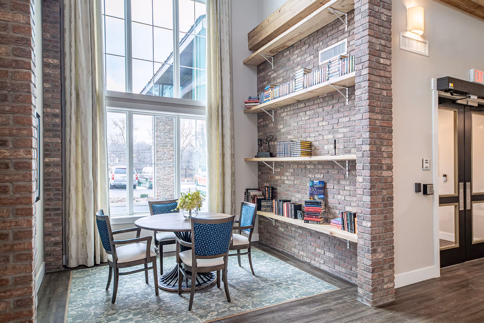 A cozy seating area with a round wooden table surrounded by four chairs with blue patterned backs and beige seats. The space features a large window with cream-colored curtains letting in natural light. A brick wall with wooden shelves filled with books is on the right side. The floor has a patterned rug, and there is a double door with glass panels in the background.