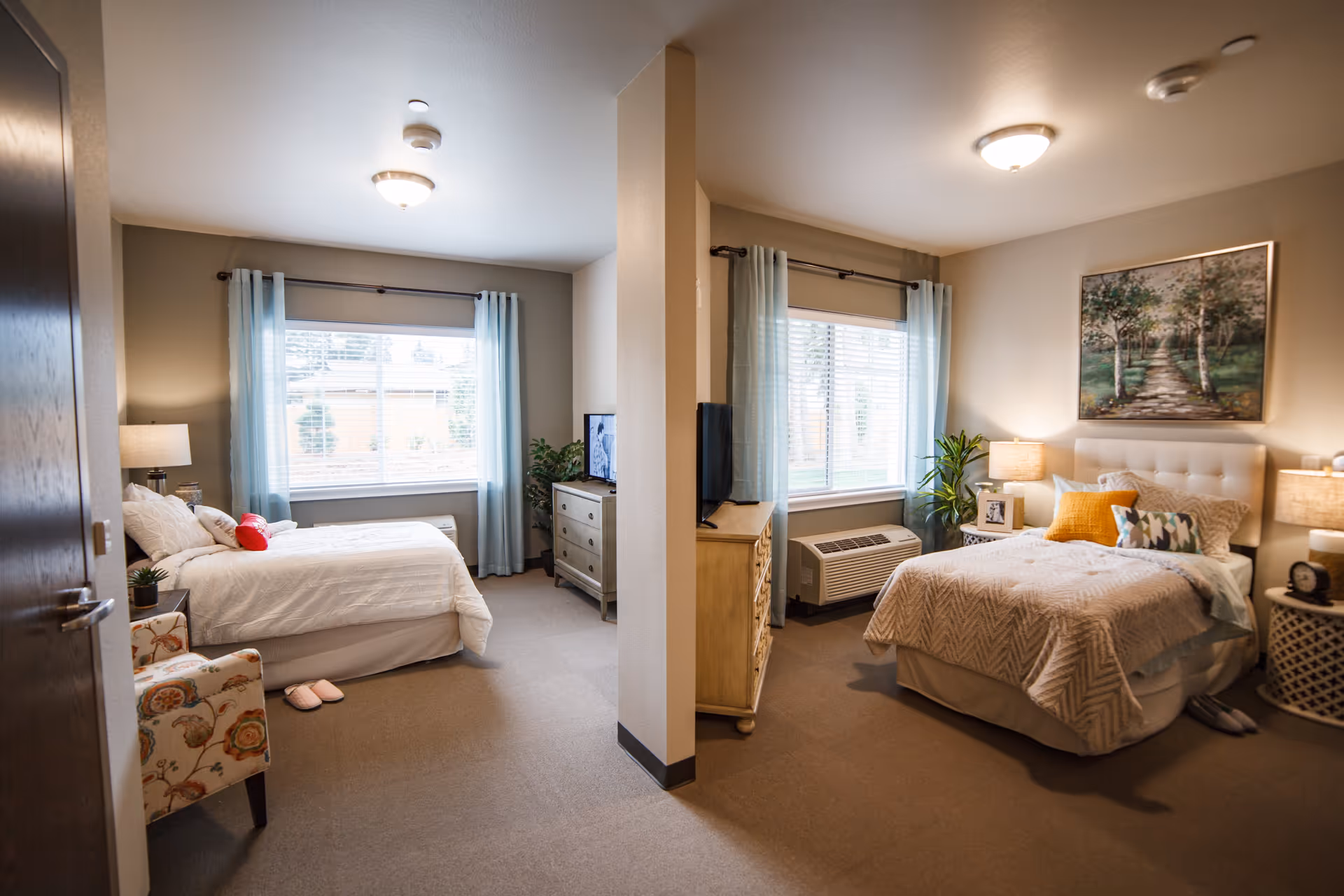 A shared bedroom in a senior living facility with two separate beds divided by a partial wall. Each side has a window with light blue curtains, a bedside table with a lamp, and a dresser with a television. The left side features a floral armchair and slippers on the floor, while the right side has a framed picture above the bed and a small plant near the window.
