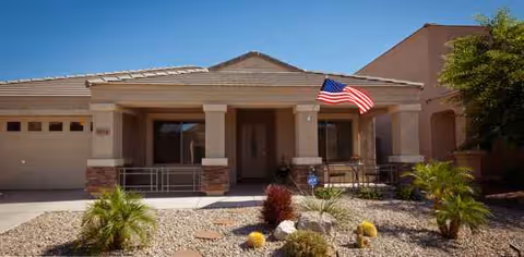 Front exterior of a single-story assisted living home with an American flag and desert landscaping.