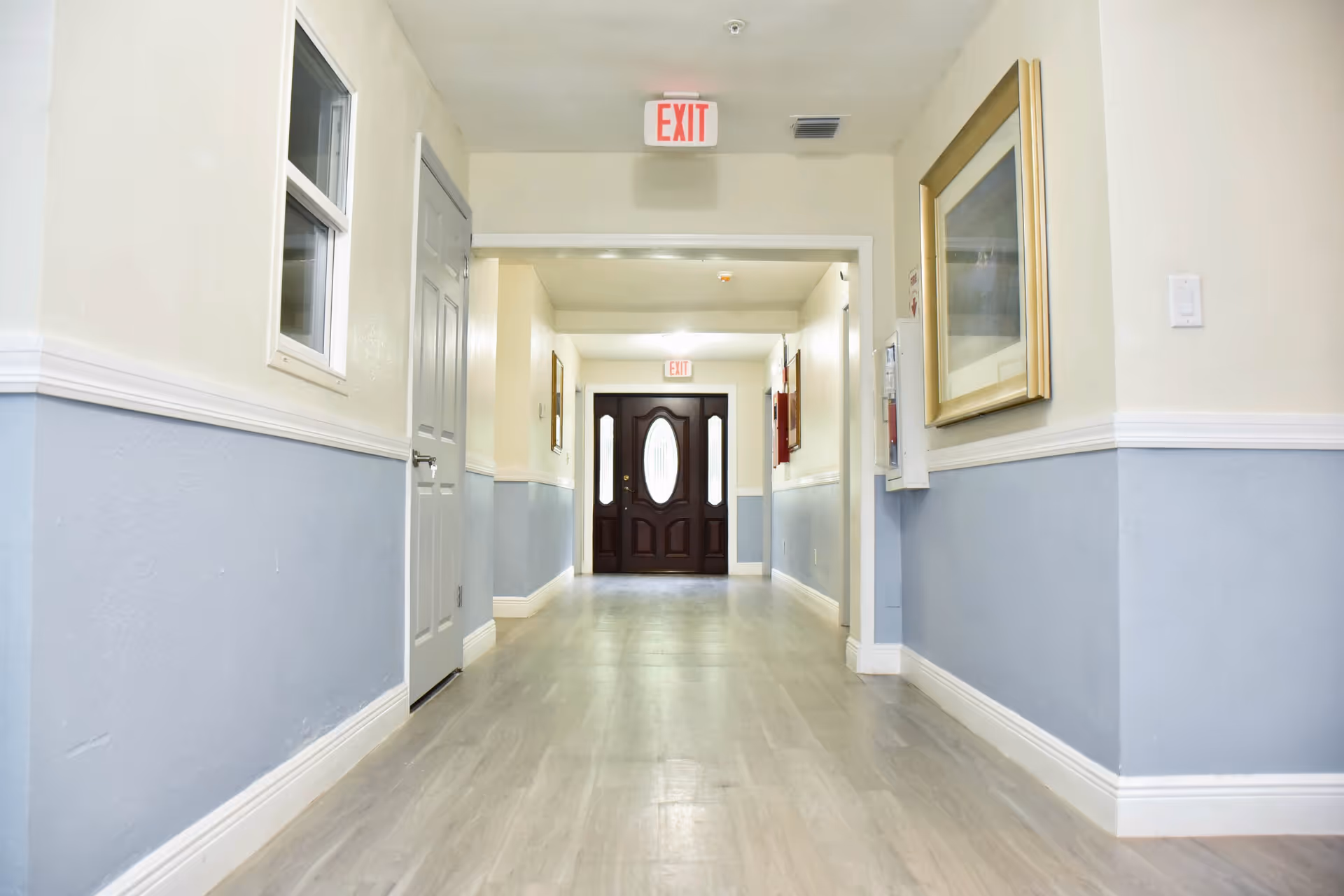 Well-lit interior hallway with blue wainscoting, wood-look flooring, exit signs, and a dark double door at the far end.