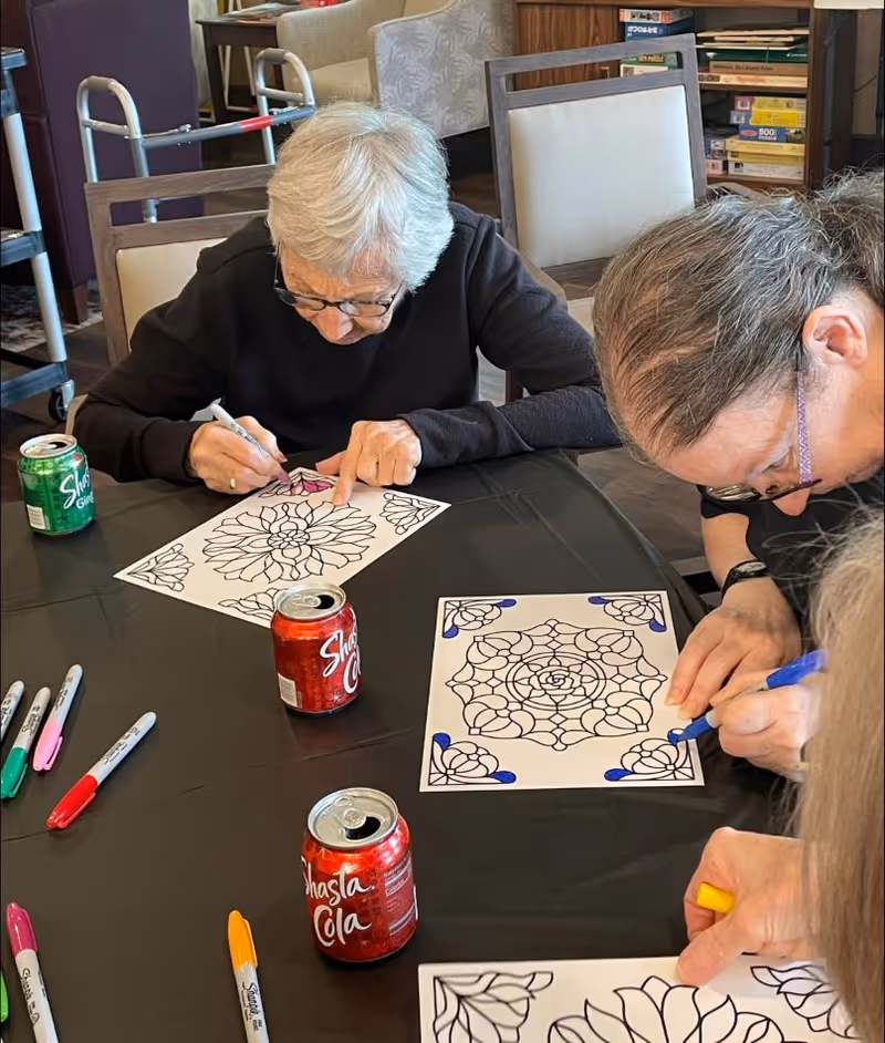 Three elderly individuals sitting around a table engaged in coloring floral mandala designs on paper using colored markers. There are several cans of Shasta Cola and other soft drinks on the table, along with various colored markers. The setting appears to be a communal indoor space with chairs and shelves in the background.