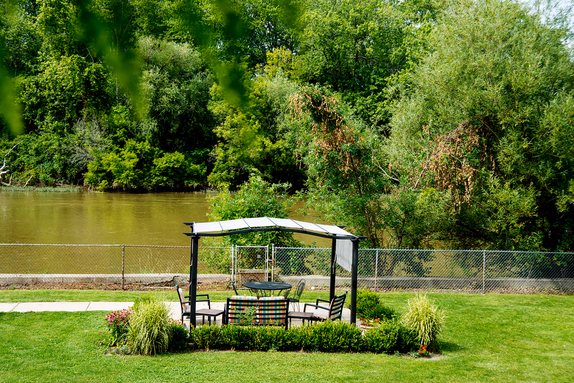 Outdoor seating area with a metal frame canopy and chairs arranged around a round table, surrounded by green grass and plants, with a river and dense trees in the background.