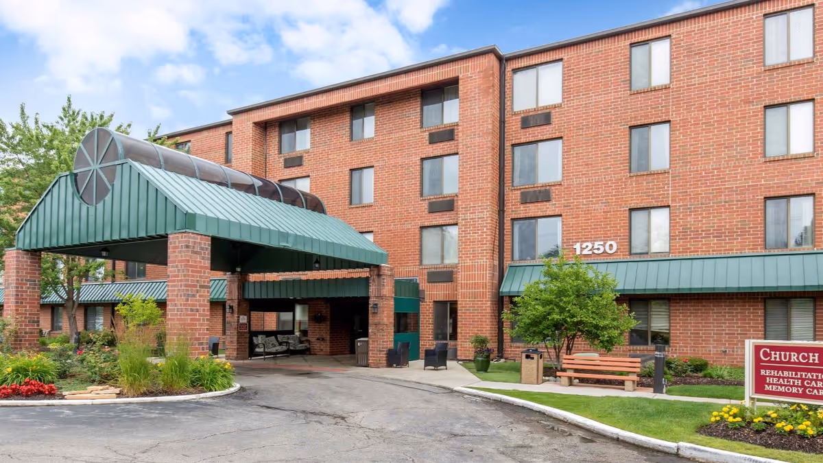 Exterior view of a multi-story brick building with a green metal awning over the entrance. The building has several windows and the number 1250 displayed near the entrance. There is a landscaped area with flowers, bushes, a tree, a bench, and a sign that reads 'Church Creek Rehabilitation Health Care Memory Care'.