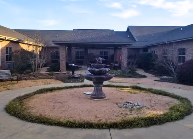 Outdoor courtyard area with a central multi-tiered fountain surrounded by a circular patch of grass and dirt. The courtyard is enclosed by a single-story brick building with multiple windows and a covered patio area with seating. There are pathways and some landscaping with bushes and small trees around the courtyard.