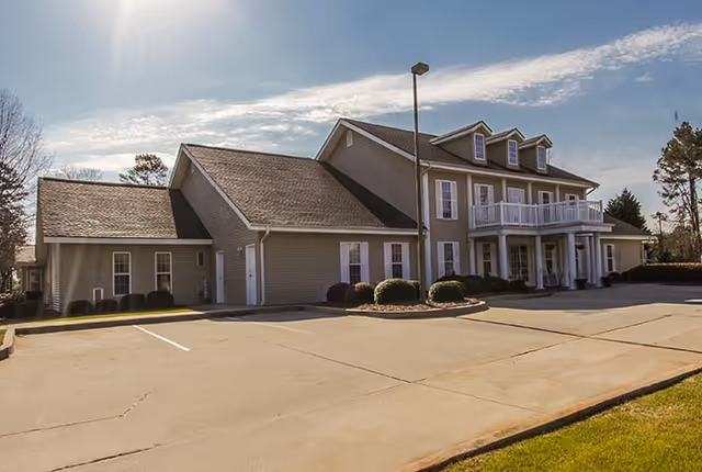 Exterior view of a two-story building with beige siding and white trim, featuring a balcony and multiple windows. The building is surrounded by a paved parking area and some landscaping with bushes. The sky is partly cloudy with sunlight illuminating the scene.